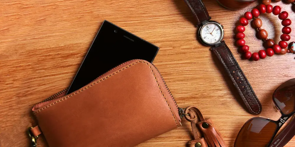 Brown leather wallet on a wooden surface with a smartphone, a leather watch strap, and a red beaded bracelet nearby.