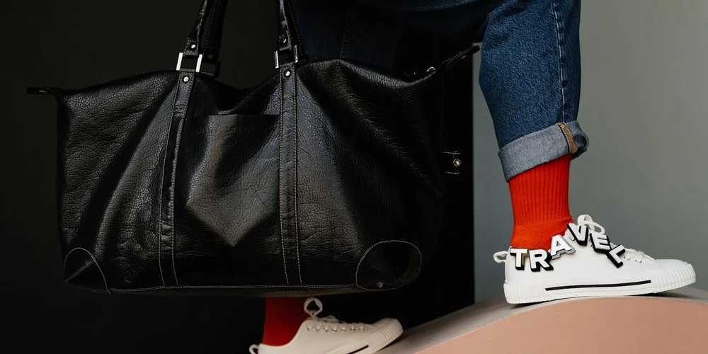 Black leather Saddleback bag with a person wearing blue jeans, red socks, and white sneakers with 'TRAVEL' on the side
