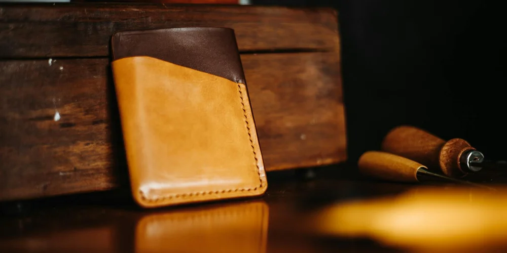 Tan leather bifold wallet resting on a wooden workbench with a leatherworking tool in the background.