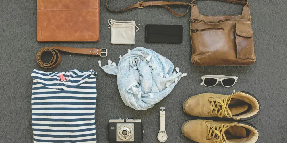 Flat lay of assorted leather items and accessories on a gray surface, including a brown leather bag, belt, shoes, a striped shirt, a scarf, sunglasses, a watch, a camera, a phone, and a small pouch.