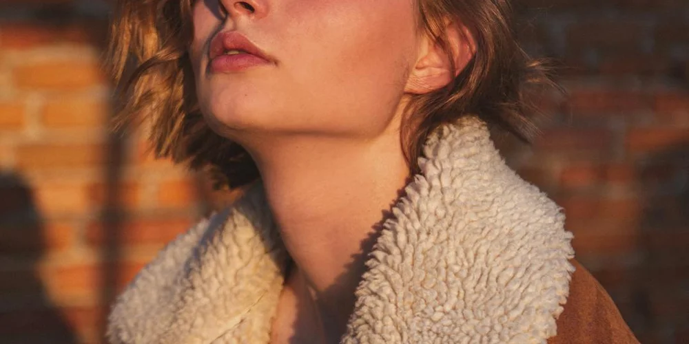 Close-up of a white fluffy sheepskin collar worn over a brown jacket with a brick wall background.