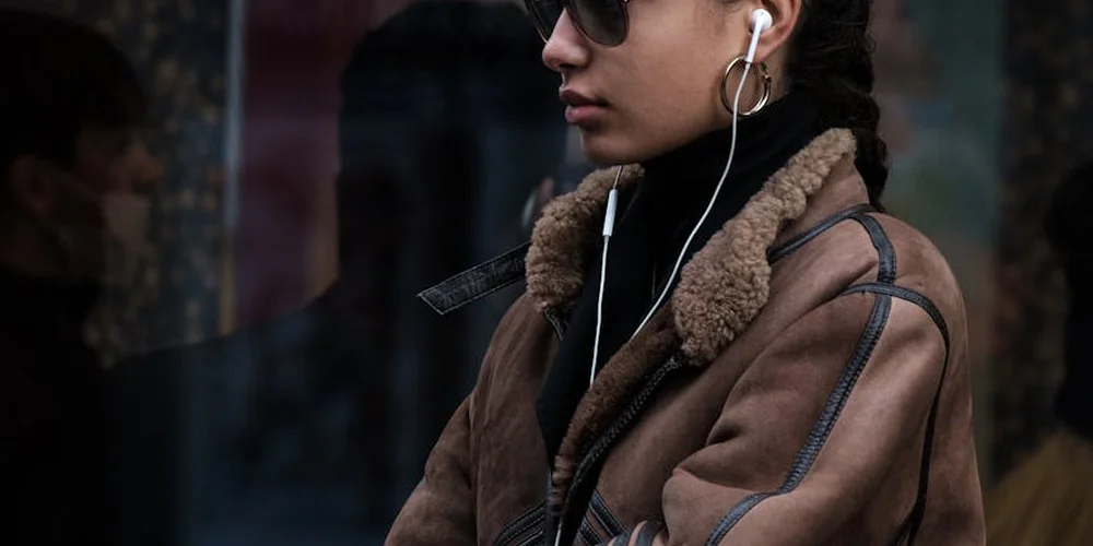 Person wearing a brown shearling leather jacket with sunglasses and earphones, outdoors.