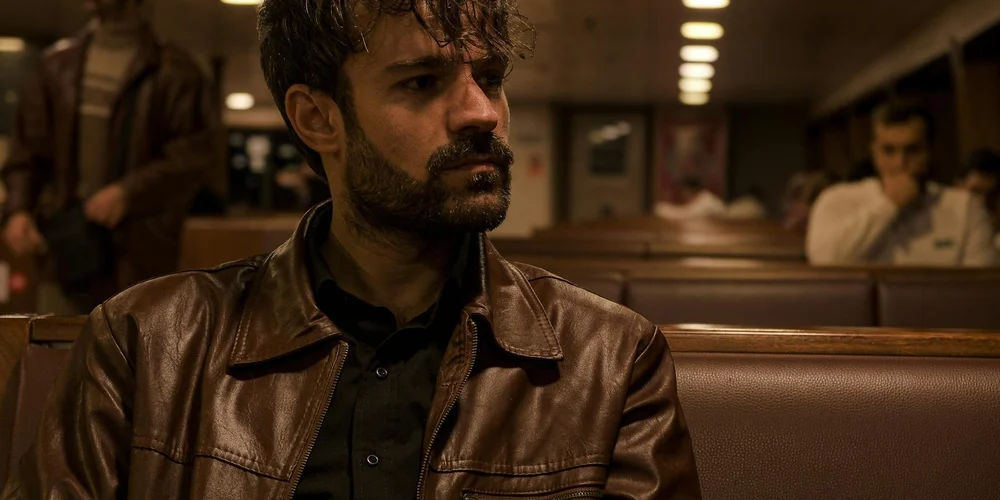 A man wearing a brown leather jacket sits on a bench in a dimly lit public space, looking to the side.