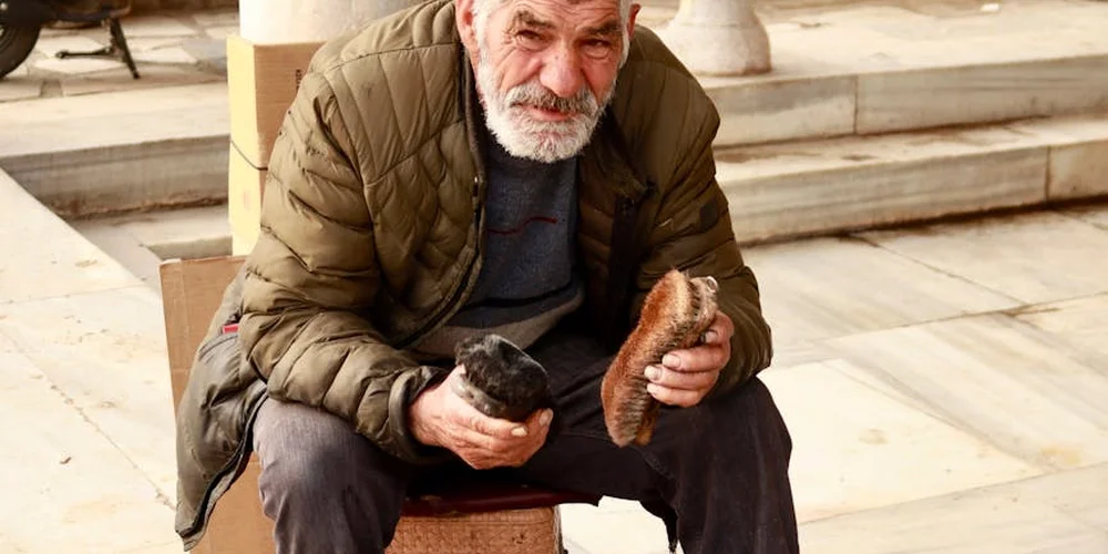 An elderly man sits on stone steps, examining two pieces of leather in his hands.