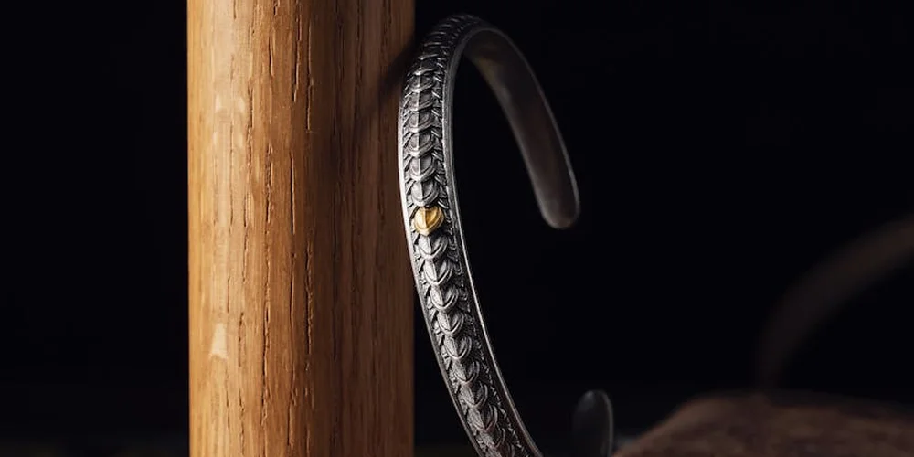 Close-up of a dark bracelet with a braided metal accent and a small gold rivet, set against a black background near a wooden post.