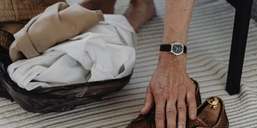 Close-up of an older person's hand with a wristwatch resting near a laundry basket and folded clothes.
