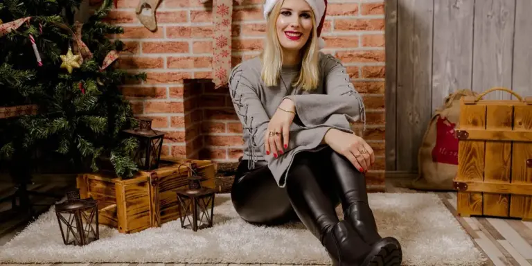 A person sits on the floor in a cozy room wearing knee-high black leather boots, with a Christmas tree and wooden crates in the background.