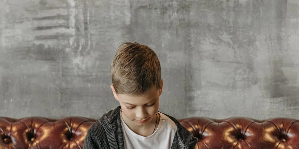 Young person with short hair looking down while seated on a brown leather couch, wearing a white shirt and dark cardigan