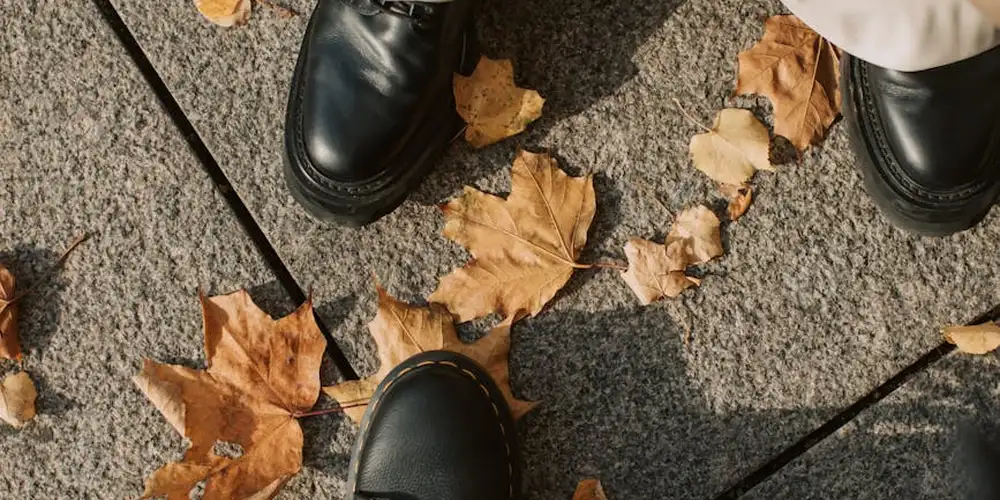 Top-down view of black leather shoes walking on a concrete sidewalk with fallen autumn leaves.