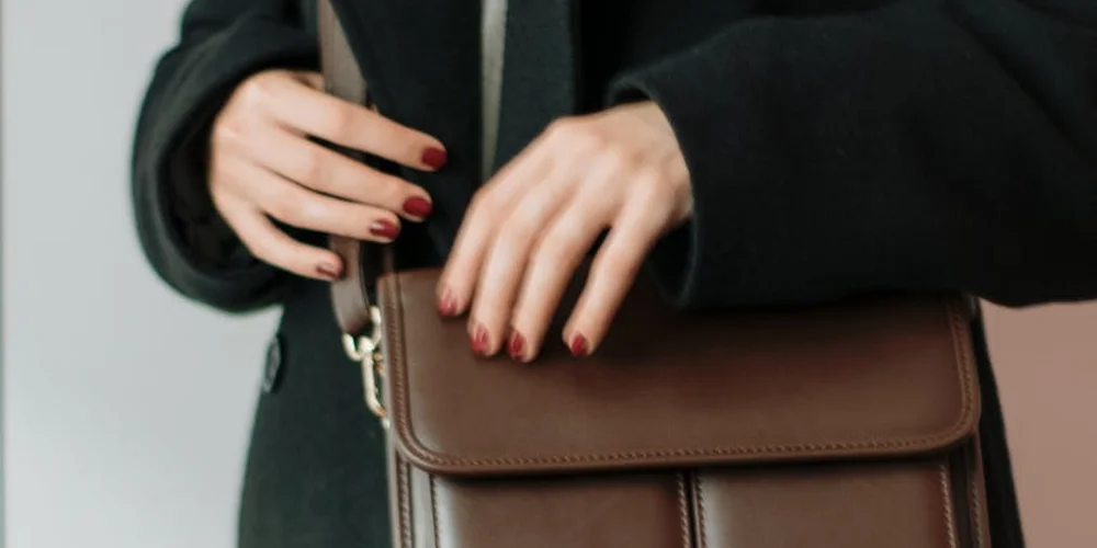 Close-up of a brown leather handbag being held by hands with red nail polish, wearing a dark coat