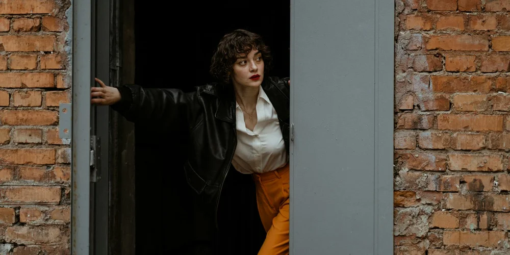 A person wearing a black leather jacket, white shirt, and orange pants steps out from a doorway between brick walls.