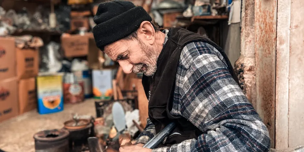 Bearded craftsman wearing a black beanie and plaid shirt repairs a leather item in a cluttered workshop.