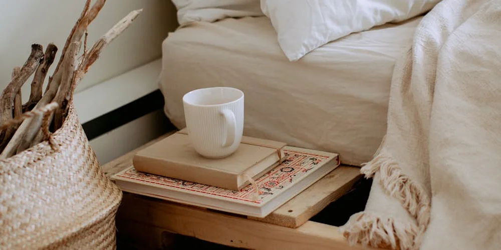 Cozy interior scene showing a mug on a stack of books on a small wooden table beside a woven basket of sticks and a beige blanket, with a soft bed in the background.
