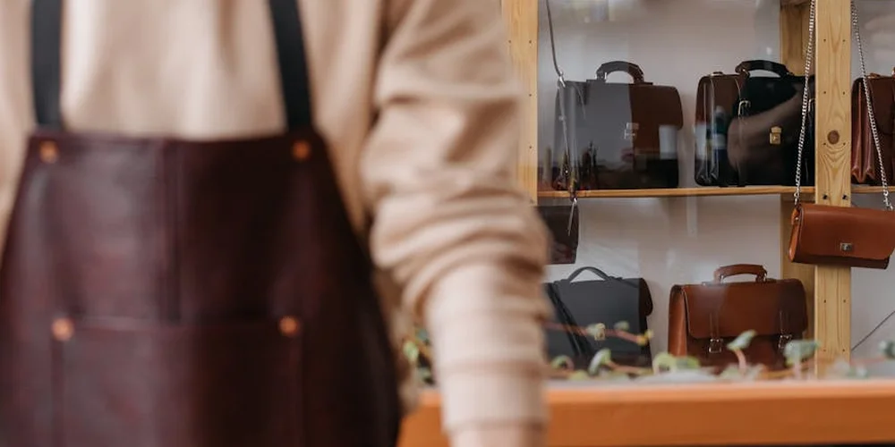 Leather bags on display shelves in a storefront, with a person in a beige sweater and apron in the foreground