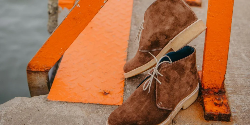 Brown suede chukka boots with light beige soles and white laces, resting on an orange railing near water