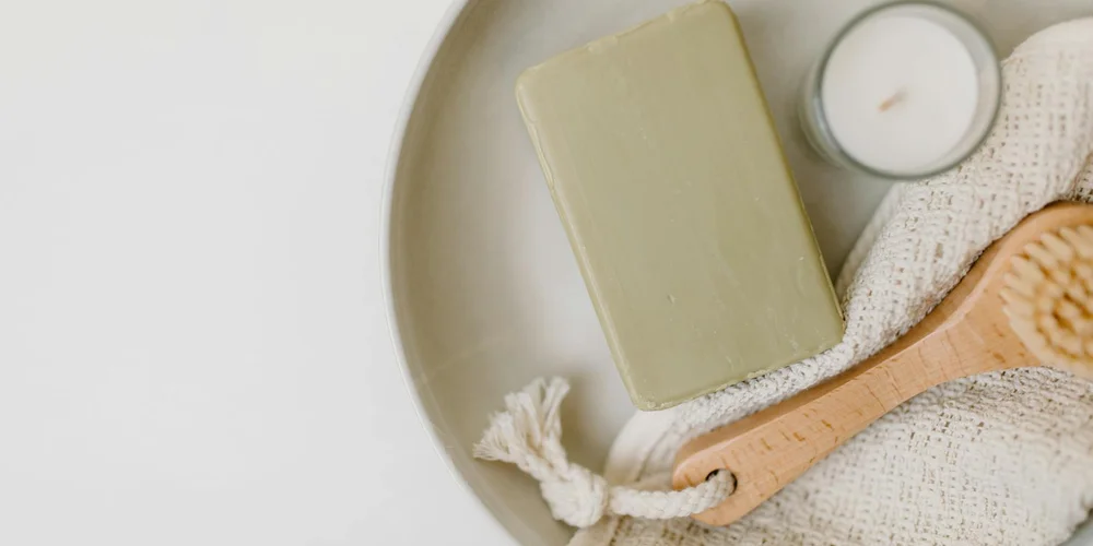 Flat lay of suede care items: a light-colored bar, a wooden-handled brush, a folded cloth, and a small white candle on a round tray.
