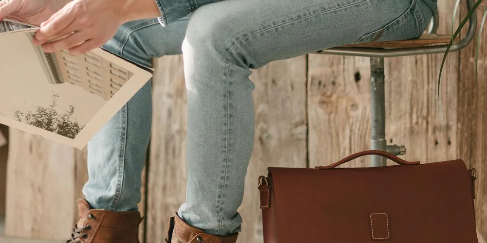 Person wearing light blue jeans sits on a wooden bench, holding a magazine. A brown leather bag rests nearby.