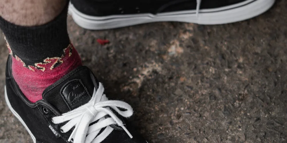 Close-up of a black suede sneaker with white laces resting on a rough pavement, next to a red sock.