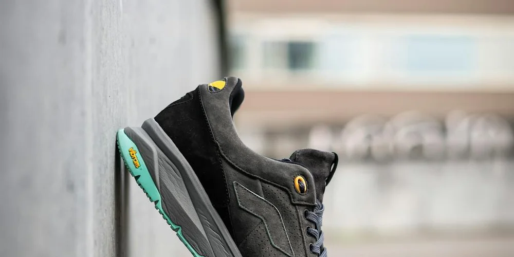 Black suede sneaker with teal sole leaning against a concrete wall.