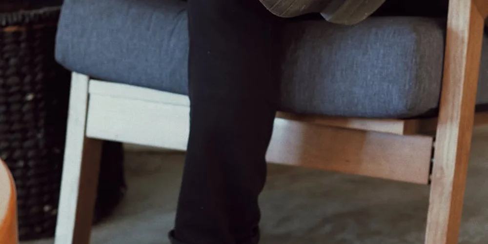 A person in dark pants sits by a blue upholstered sofa with a wooden frame, set in a tidy indoor space.