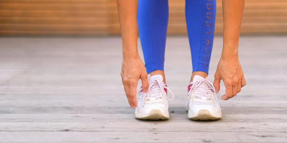Close-up of a person tying white sneakers on a light floor, wearing blue leggings.