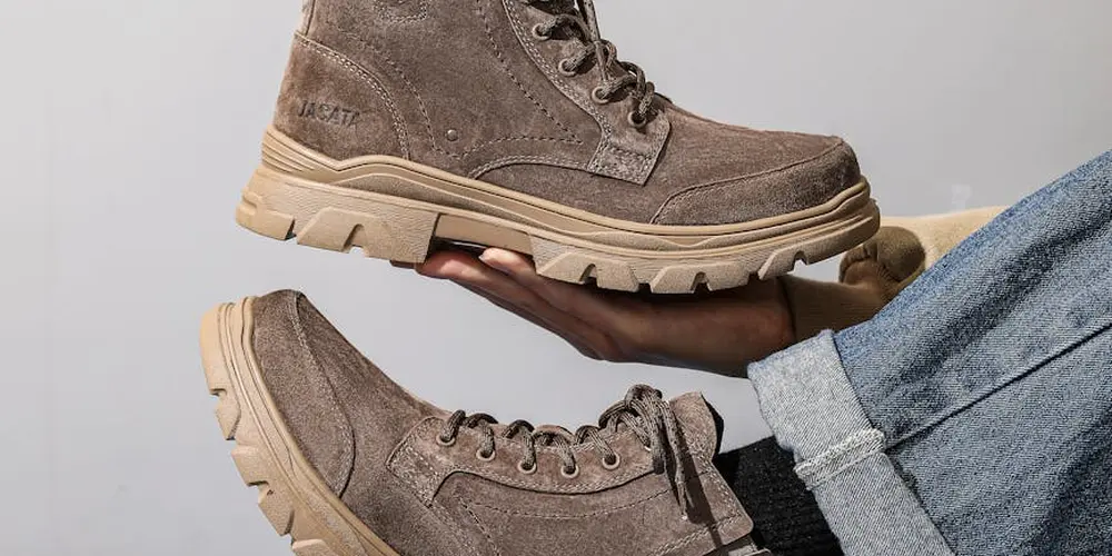 Two brown suede hiking boots with thick beige soles; one boot is being held in a hand while the other rests on the floor.