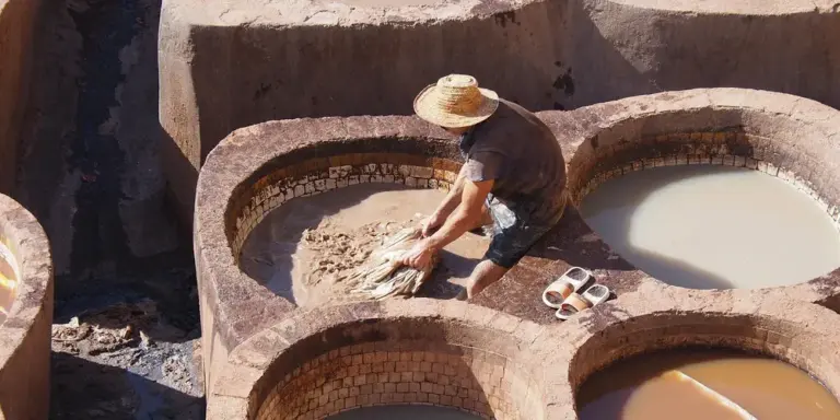 Tanner wearing a wide-brimmed hat kneads hides in circular stone tanning vats filled with liquid.