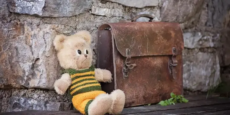 A plush teddy bear sits beside a worn leather bag against a stone wall, illustrating leather care and stain removal.