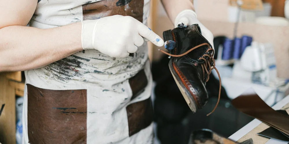 Cobbler wearing gloves examining a leather dress shoe and using a tool to smooth creases in a workshop.