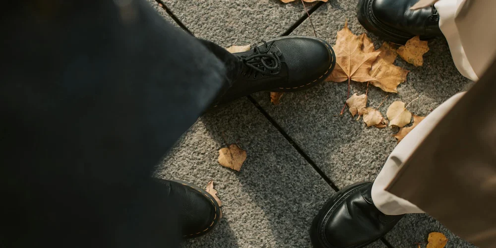 Feet in black leather shoes on a sidewalk with autumn leaves, suggesting a setup for adhesive removal.
