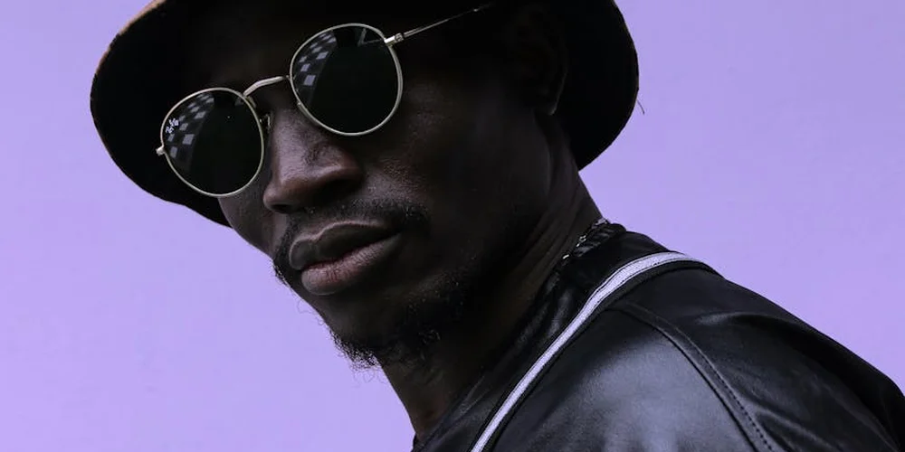 Portrait of a person wearing a black leather jacket, round sunglasses, and a hat against a purple backdrop