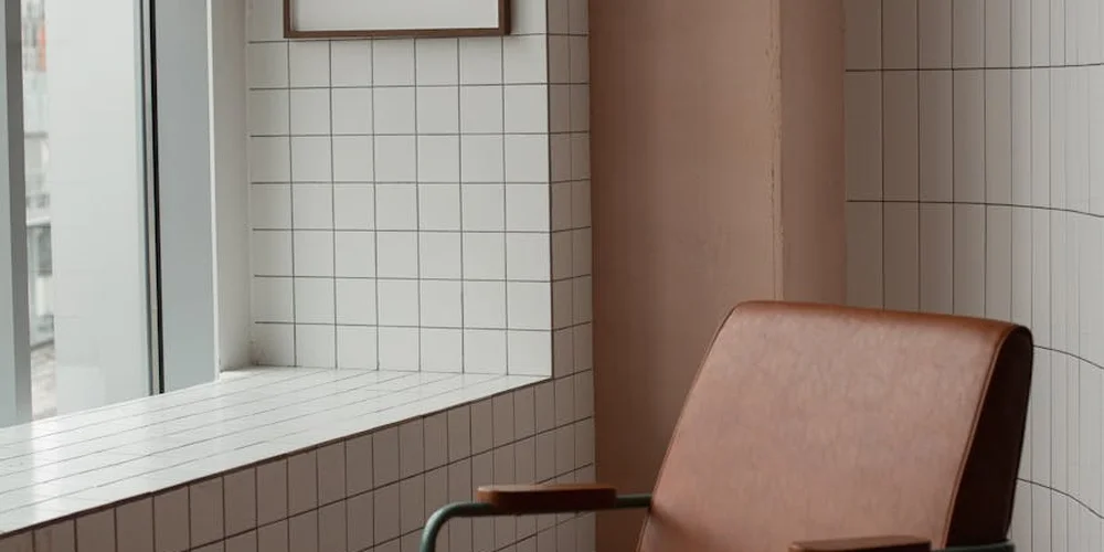 Brown leather chair beside a window with white tiled walls in a bright room.