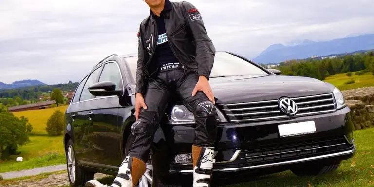 Person wearing black leather motorcycle jacket and pants stands beside a dark Volkswagen car in a rural landscape.