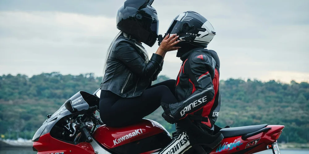 Two motorcyclists wearing black and red leather gear with full-face helmets sit on a red Kawasaki motorcycle, adjusting helmets.