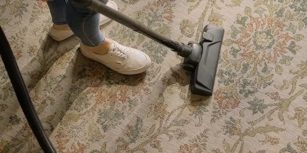 Person vacuuming a patterned rug on a beige floor with a handheld vacuum