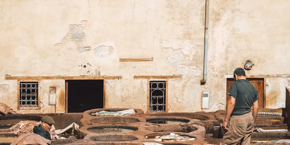 Two workers in a tannery yard examine hides arranged around circular vats used for vegetable tanning, with a weathered wall and small windows in the background.