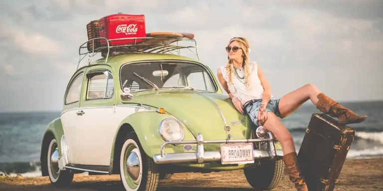 A woman sits on the front bumper of a light-green vintage car by the seaside, with luggage on the roof rack, under a partly cloudy sky.