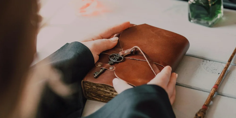 Hands resting on a brown leather-bound journal with a string and key pendant on a white wooden table.