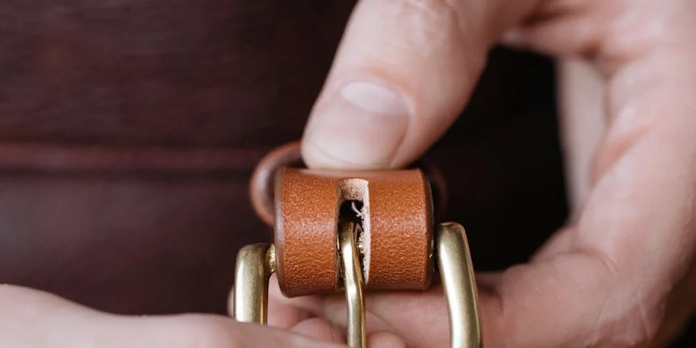 Close-up of a hand holding a small brown leather strap with a torn edge and brass hardware, against a dark background.