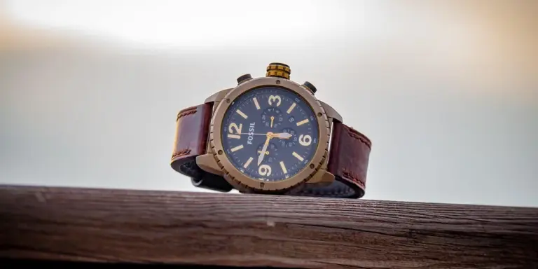 Vintage wristwatch with a brown leather strap and brass case, resting on a weathered wooden railing outdoors.