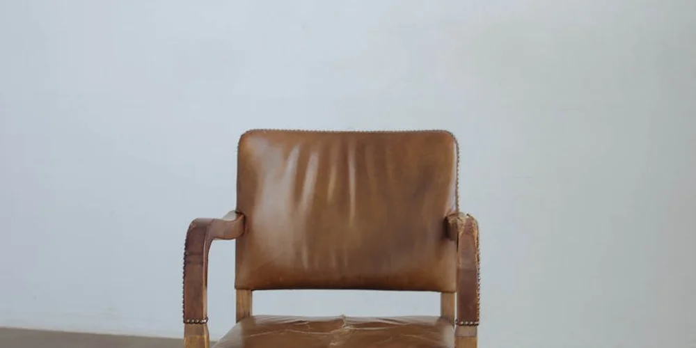 Brown leather armchair with a wooden frame and nailhead trim, set against a pale wall.