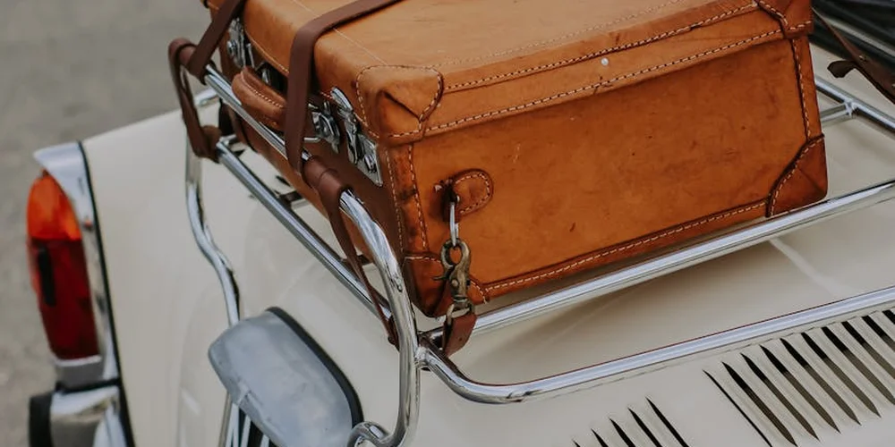 A close-up of a weathered brown leather suitcase strapped to the chrome luggage rack on the back of a white vintage car.