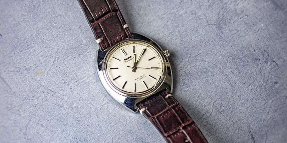 Close-up of a luxury-style wristwatch with a brown leather strap on a textured gray surface.
