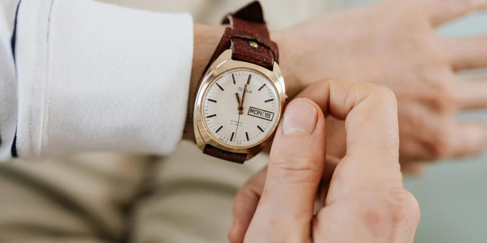 Close-up of a brown leather watch strap being adjusted on a wearer's wrist, with a white shirt sleeve visible