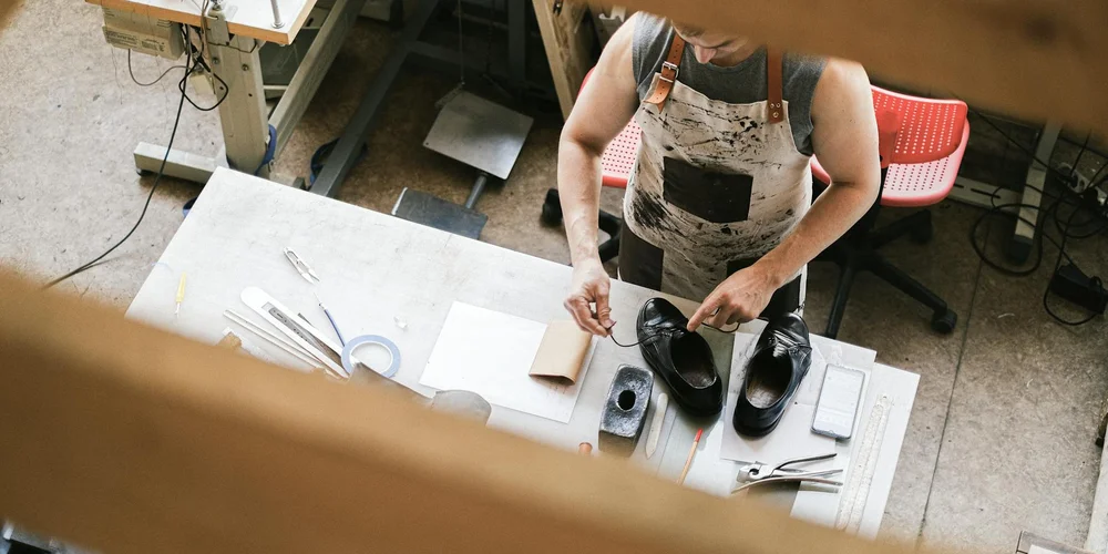 A craftsman in a workshop repairs cracked leather shoes at a workbench, with tools and leather pieces laid out.