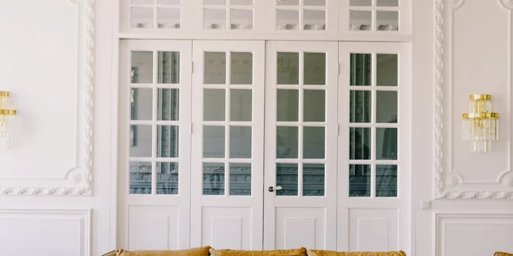 Two white double doors with multiple glass panes, ornate wall paneling, and gold wall sconces on either side.