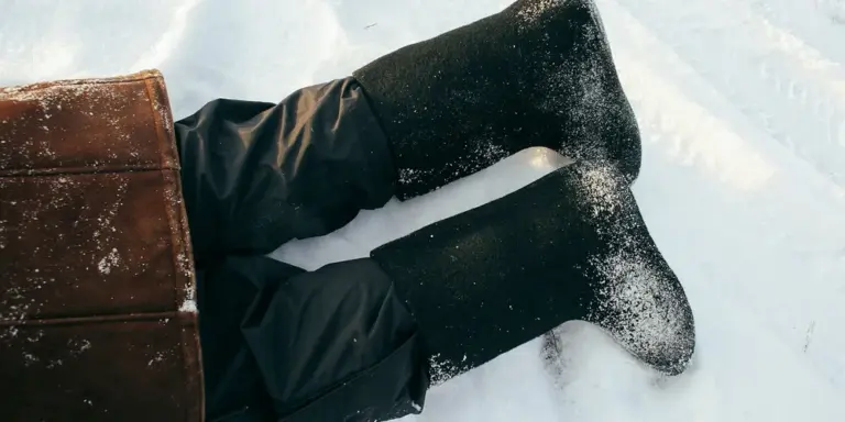 A pair of black leather winter boots on snowy ground, with snow clinging to the soles and pant legs tucked in.