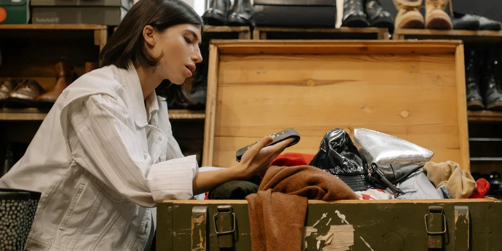 A woman in a light shirt examines leather items inside an open wooden crate in a shop.