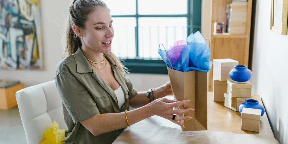 A woman sits at a desk, placing blue and purple tissue paper into a brown paper gift bag.