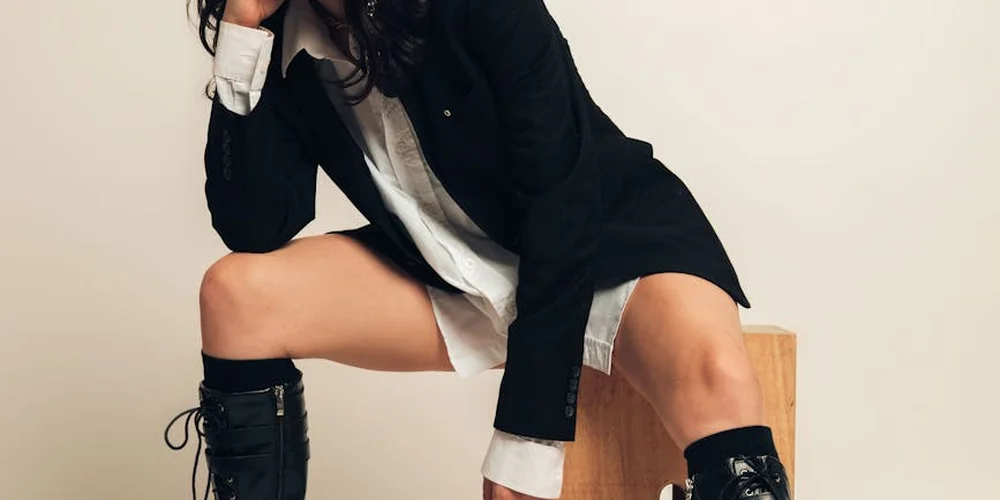 Person wearing black suede knee-high boots sits on a wooden block, dressed in a black blazer and white shirt.